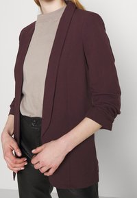 Nainen pukeutuneena viininpunaiseen bleiseriin, jossa on rypytetyt hihat, beigen värisen korkeakauluksisen topin ja mustien korkeavyötäröisten housujen kanssa, kädet ristissä vyötäröllä.