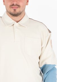 Polo shirt met lange mouwen in gebroken wit met lichtblauwe mouwen, subtiele bruine accenten, een borstzak en een ronde badge op de mouw.