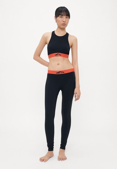 Zwarte sportieve crop top met een oranje band en logo, gecombineerd met zwarte legging met een bijpassende oranje tailleband. Glad, rekbaar materiaal.
