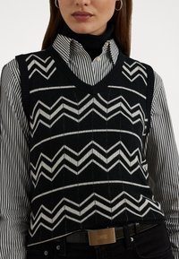 Czarnobiała dzianinowa kamizelka w wzór jodełki noszona na paskowanej koszuli na guziki, z dekoltem w serek i prążkowanymi krawędziami, z detalem w postaci paska.