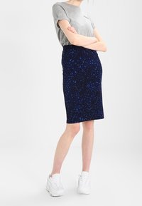 Grå t-shirt med logotyp, tillsammans med en mörkblå leopardmönstrad pennkjol. Modellen bär vita sneakers, har armarna i kors, i en neutral miljö.