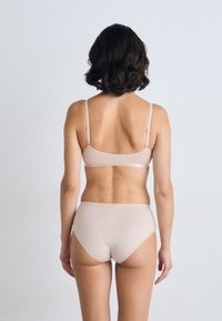 Vrouw met kort donker haar, gekleed in een beige bh en een hoge taille onderbroek, staand met de rug naar de camera tegen een effen lichte achtergrond.