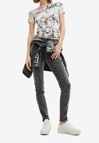 Camiseta con estampado de graffiti en blanco, jeans skinny grises con bandas laterales y zapatillas blancas. Chaqueta de mezclilla adicional atada a la cintura.