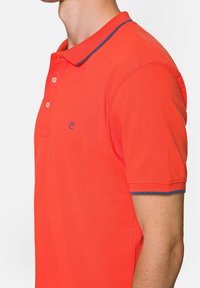 Czerwona koszulka polo z klasycznym kołnierzykiem, z kontrastowym niebieskim wykończeniem na krawędzi. Zapięcie na trzy guziki i logo wyszywane na piersi.