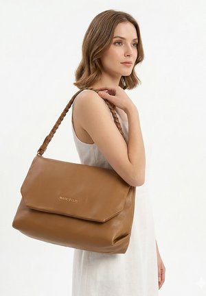 Frau in weißem Kleid trägt große braune Marc Ellis Schultertasche mit geflochtenem Riemen vor einfachem Hintergrund.