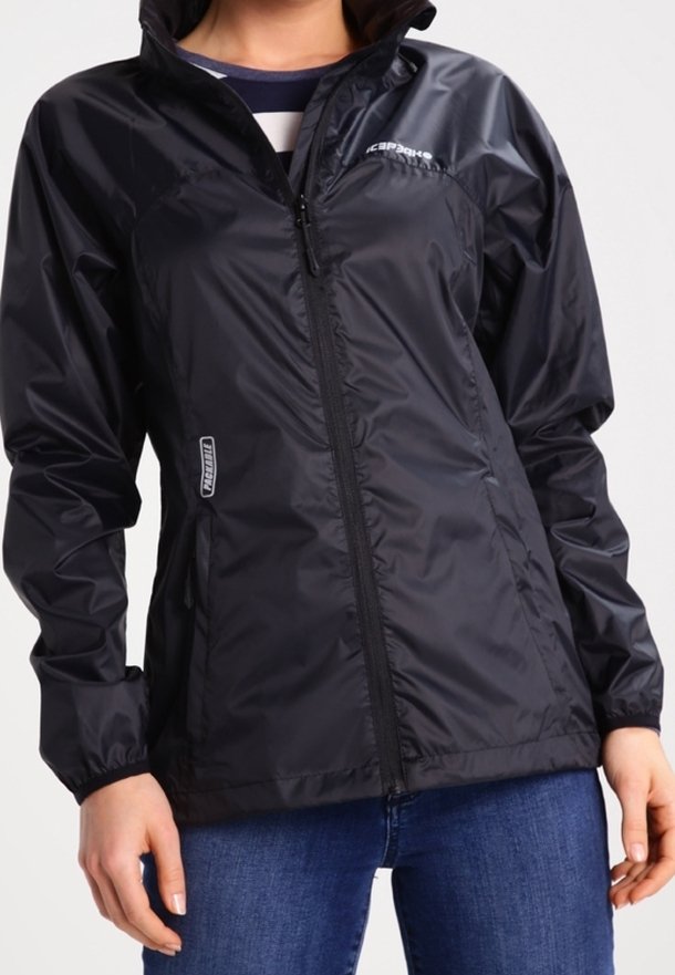 Personne portant une veste de pluie légère noire zippée avec des poignets élastiques et un jean bleu, debout devant un fond uni.