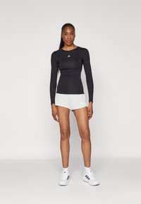 Haut de sport noir à manches longues avec un col rond, associé à un short gris clair. Des baskets blanches complètent la tenue, arborant des logos de marque.