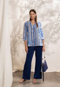 Blouse à motif bleu avec manches trois-quarts, associée à un pantalon large bleu marine ; le mannequin tient un sac à main bicolore avec une bandoulière.