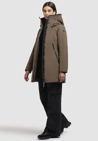 Hnedý zateplený parka kabát s vysokým golierom, predným zipsom a dvomi bočnými vreckami. Nosený cez čierny outfit, doplnený čiernymi cargo nohavicami.