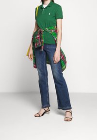 Grön polo skjorta, blå jeans, rutigt skjorta knuten i midjan, svarta sandaler med remmar och genomskinlig klack. Avslappnad outfit med olika texturer och mönster.