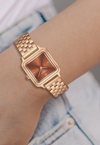 Roosakuldne ruudukujuline kell pruunikate dialiga, metallkett ja lihtsad tunni markerid. Korpusel on sile viimistlus ja minimalistlik disain.