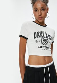Valkoinen ribattuja crop-top mustilla yksityiskohdilla, jossa on graafinen printti, joka sisältää tekstin "University of Oakland" ja laakerikuvion.
