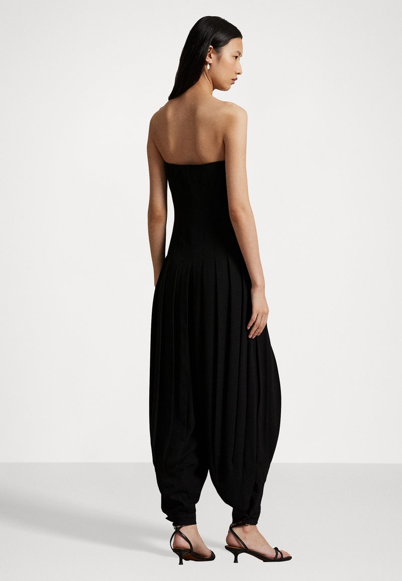 Donna in tuta nera senza spalline con pantaloni svasati e sandali neri a tacco basso con cinghie, in piedi davanti a uno sfondo bianco semplice.