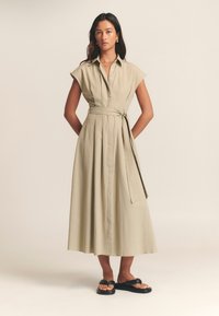 Haljina boje maslinasto zelene, s gumbima, kratkih rukava, s pojasom u struku i A-kroja. Glatke teksture i do koljena dužine. Crne sandale.