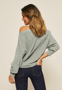 Maglione grigio chiaro a coste con design off-shoulder, maniche lunghe e vestibilità rilassata, abbinato a jeans attillati blu scuro.