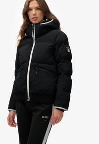 Chaqueta de plumas negra con dos bolsillos frontales con cremallera, un cuello alto y una capucha. Con detalles de acento en blanco y puños acanalados.