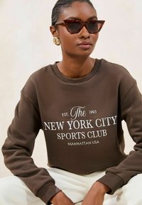 Felpa marrone con testo bianco che dice "NEW YORK CITY SPORTS CLUB" e "EST. 1993". Modello con occhiali da sole a occhio di gatto e grandi orecchini dorati.