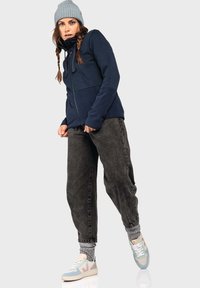 Navy-Fleecejacke mit hohem Kragen, Reißverschluss vorne und zwei Seitentaschen, kombiniert mit lockeren grauen Stoffhosen und pastellfarbenen Sneakers.