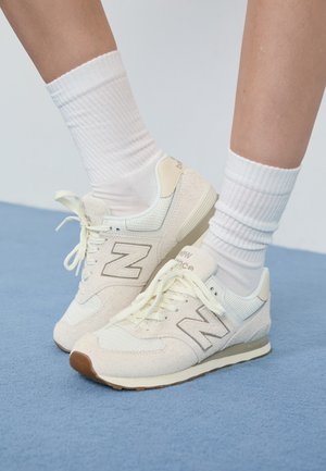 Ben iført hvide ribstrikkede crew-sokker og off-white New Balance-sneakers stående på en blå tekstureret gulv.