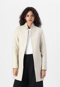 Cappotto in misto lana beige con taglio dritto, colletto alzato e tasche laterali, indossato sopra un top bianco e pantaloni neri.