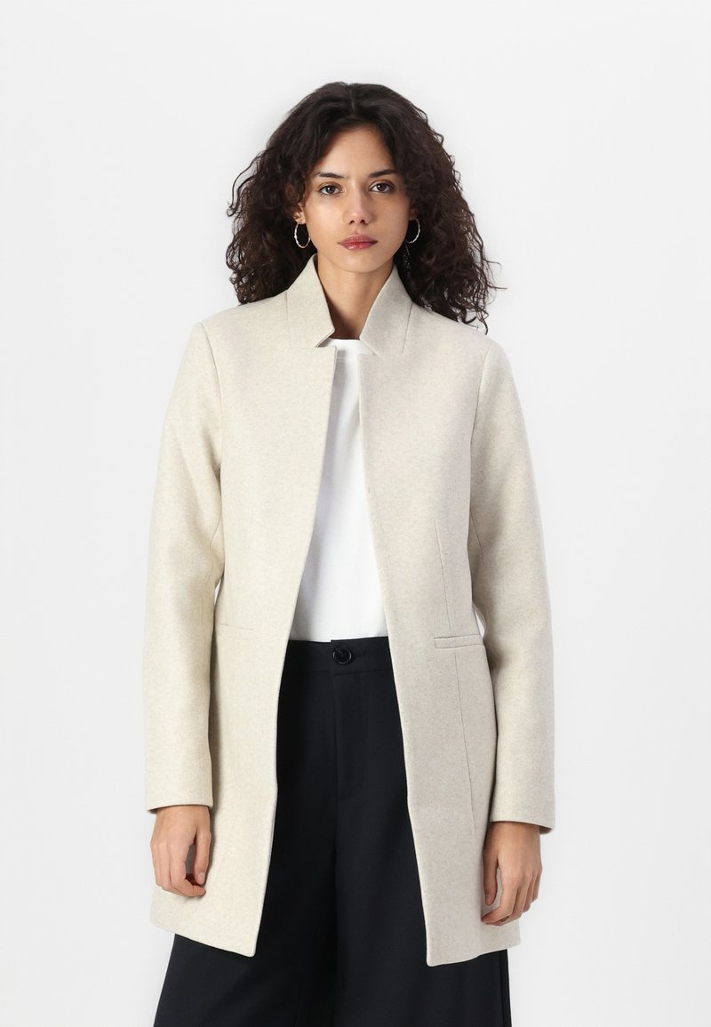 Cappotto in misto lana beige con taglio dritto, colletto alzato e tasche laterali, indossato sopra un top bianco e pantaloni neri.