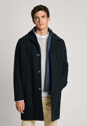 Cappotto in lana scura con colletto classico, chiusura a bottoni e tasche laterali, indossato sopra un maglione a coste grigio chiaro.