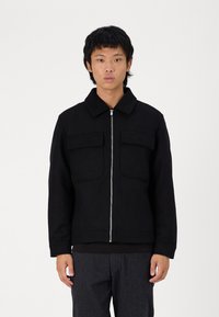 Veste zippée noire avec un col, dotée de deux poches poitrine, fabriquée dans un tissu épais et à coupe droite. Associée à un pantalon texturé gris.