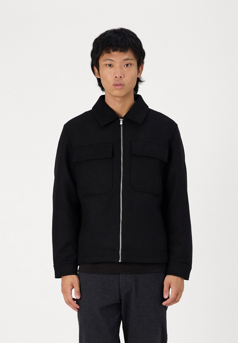 Veste zippée noire avec un col, dotée de deux poches poitrine, fabriquée dans un tissu épais et à coupe droite. Associée à un pantalon texturé gris.