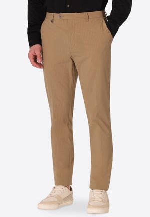 Uomo che indossa pantaloni beige a righe sottili con tasche anteriori e sneakers beige, in piedi con una mano in tasca e camicia nera.