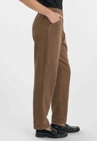 Persona que lleva pantalones marrones texturizados con bolsillos laterales y mocasines negros brillantes, de pie contra un fondo claro y liso.