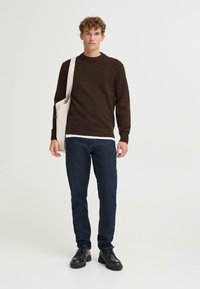 Brązowy sweter z dzianiny, ciemne dżinsy i czarne skórzane buty. Modelka trzyma jasnokolorową torbę z płótna. Prosty, codzienny strój.