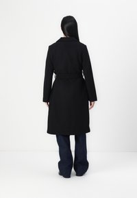 Manteau en laine noir avec une taille ceinturée, des manches longues et un col, associé à un pantalon en denim à jambes larges. Texture lisse, design mi-long.