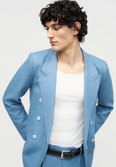 Jeune personne aux cheveux courts et bouclés portant une veste de costume bleu clair, un débardeur blanc et une ceinture noire, regardant vers la gauche avec les mains dans le dos.