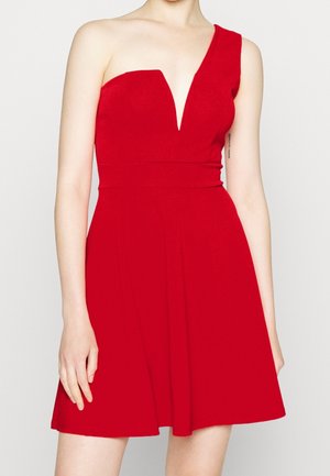 Kvinde iført en ærmeløs, knaldrød kjole med asymmetrisk halsudskæring og udvidet nederdel, stående mod en ensfarvet lys baggrund.