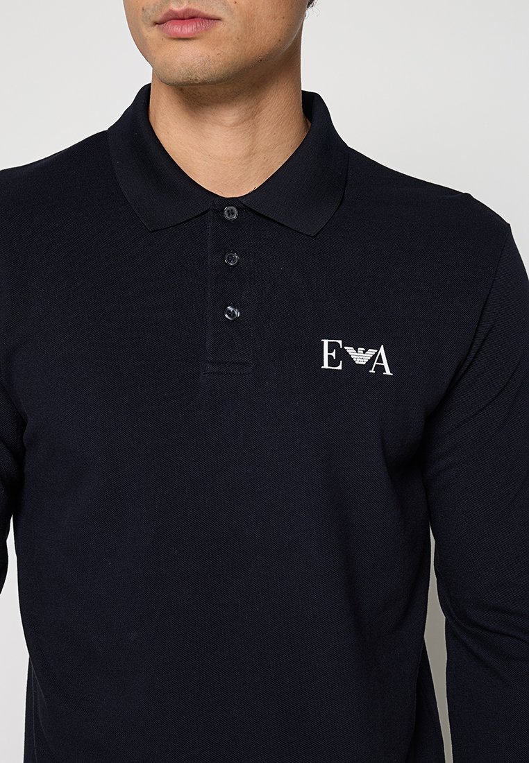 Hombre que lleva una camiseta polo negra de manga larga con tres botones y un logo blanco bordado en el pecho izquierdo.