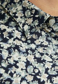 Chemise florale avec un fond bleu marine foncé et des fleurs vert clair et crème. Conception à col avec un détail de bouton unique. Tissu doux au toucher.