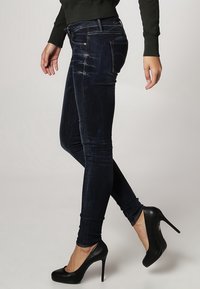 Jeans ajustados de color azul oscuro, hechos de una tela suave y lisa. Presentan un diseño clásico de cinco bolsillos y un ligero desgaste, combinados con tacones altos negros.