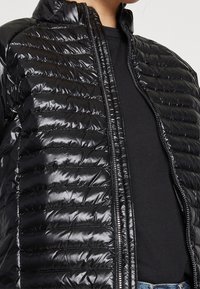 Svart, kviltad pufferjacka med glänsande yta, hög krage och hel dragkedja. Den har horisontella sömmar och en lätt design.