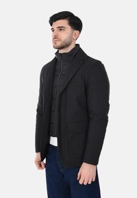 Blazer nero con finitura lucida, dotato di chiusura a due bottoni, tasche laterali e indossato sopra una giacca trapuntata con collo alto.