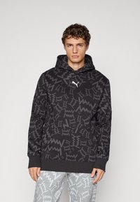 Ung mand iført sort Puma hoodie med abstrakt grå zigzag-mønster og matchende grå bukser med et lignende mønster, stående foran en ensfarvet baggrund.