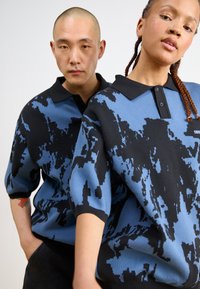 Πλεκτά πόλο μπλουζάκια σε μπλε και μαύρο αφηρημένο σχέδιο, με μαύρο γιακά και κοντά μανίκια, φορεμένα από δύο μοντέλα σε ουδέτερη φόντο.