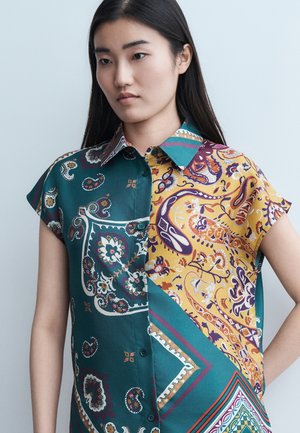 Mujer que lleva una camisa de manga corta abotonada con patrones de cachemira contrastantes en tonos turquesa y amarillo, de pie frente a un fondo claro y liso.