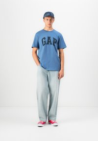 Tânăr îmbrăcat cu un tricou albastru GAP, blugi lejerați de culoare albastru deschis, adidași roșii și o șapcă albastră, stând în fața unui fundal alb.