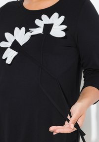 Abito nero con applicazioni di fiori bianchi e dettaglio arricciato con una lunga tracolla nera, che mette in risalto una texture liscia.