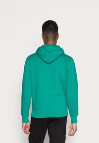 Vihreä hupullinen collegepaita takaapäin nähtynä, jossa on yksinkertainen design, pehmeä pinta ja resoriset hihansuut. Yhdistetty mustiin alaosiin.