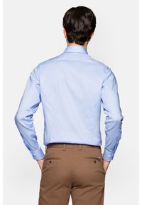 Jasnoniebieska koszula z długimi rękawami, z wyraźnie widocznymi guzikami, zestawiona z brązowymi spodniami, pokazana z tyłu.