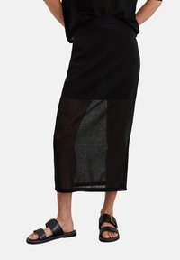 Schwarzer Midirock mit einem durchsichtigen unteren Abschnitt, der eine glatte Stofftextur aufweist. Kombiniert mit flachen schwarzen Sandalen mit Schnallen.