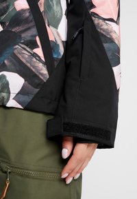 Main avec vernis blanc ajustant le poignet noir d'une veste à motif floral sur un pantalon vert olive avec poche zippée.
