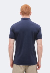 Hombre con cabello corto y rojo que lleva una camisa polo azul marino de manga corta y pantalones cortos beige, visto desde atrás sobre un fondo liso.