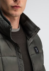 Dunkelgrüne Pufferjacke mit gestepptem Design und hohem Kragen. Darunter wird ein strukturierter brauner Pullover getragen; Logo-Patch auf der Jacke.